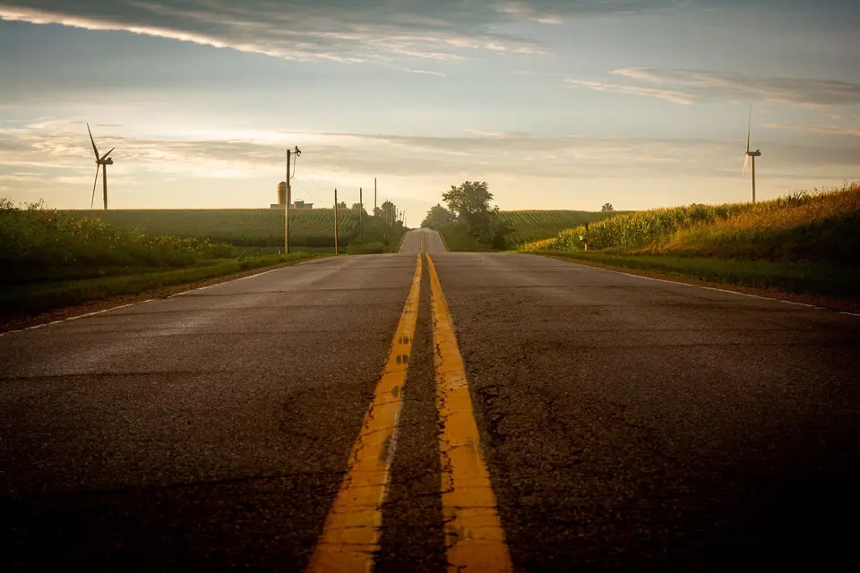 Two-lane country road with a double yellow centre line running between cornfields towards the horizon at sunrise