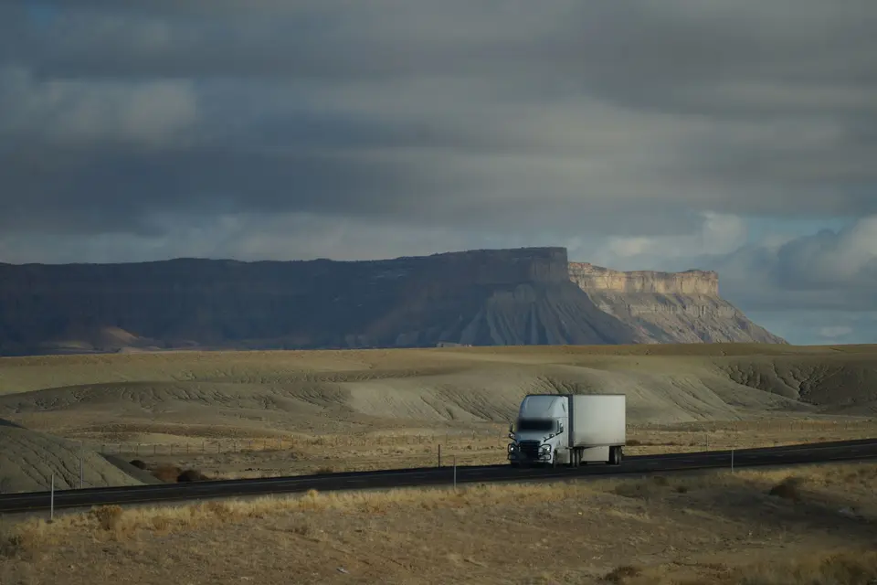 White semi-truck with a plain trailer on a highway crossing dry open country below a long mesa